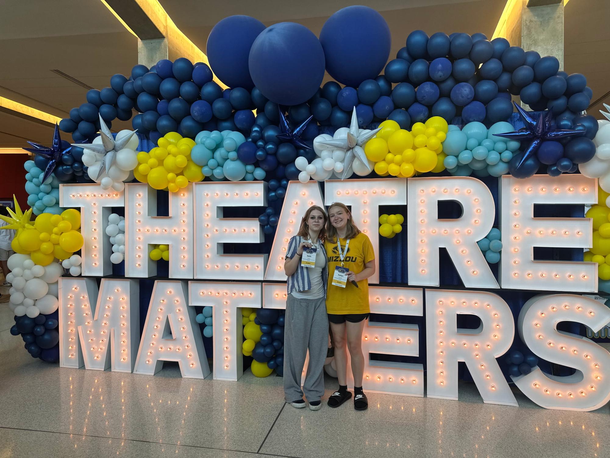 Stella Kolb (11) and Kaitlyn Bridges (12) pose in front of a Theatre Matters sign at the ITF opening ceremony. They rehearsed for hours preparing their performance. They scored an Excellent at the national level.